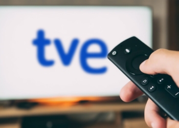 Una mano sostiene un mando a distancia frente a una pantalla de televisión con el logotipo de TVE desenfocado.