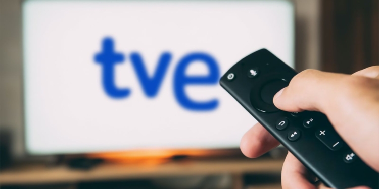 Una mano sostiene un mando a distancia frente a una pantalla de televisión con el logotipo de TVE desenfocado.