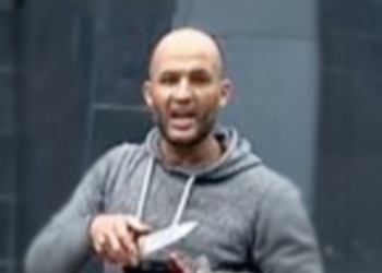 Hombre con sudadera gris sostiene un objeto en la mano en un entorno urbano desenfocado.