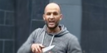 Hombre con sudadera gris sostiene un objeto en la mano en un entorno urbano desenfocado.