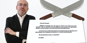Hombre con traje y brazos cruzados junto a dos machetes cruzados y una carta mostrada en primer plano.