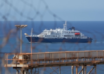 Hantavirus: Crucero de tamaño medio navegando cerca de la costa, visto desde una estructura portuaria en primer plano.