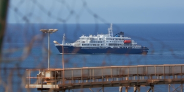 Hantavirus: Crucero de tamaño medio navegando cerca de la costa, visto desde una estructura portuaria en primer plano.