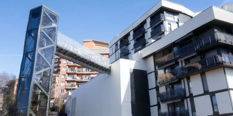 Lugar donde Un magrebí reincidente fracturó la clavícula a una anciana e intentó ahogar a su marido en Bilbao. Edificio residencial moderno con una pasarela elevada conectada a una torre metálica exterior.