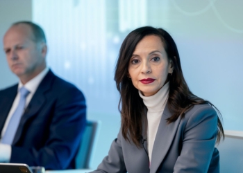¿607 millones más? El nuevo sablazo en tu factura de luz. Beatriz Corredor con traje gris en una reunión mientras un hombre aparece desenfocado al fondo.