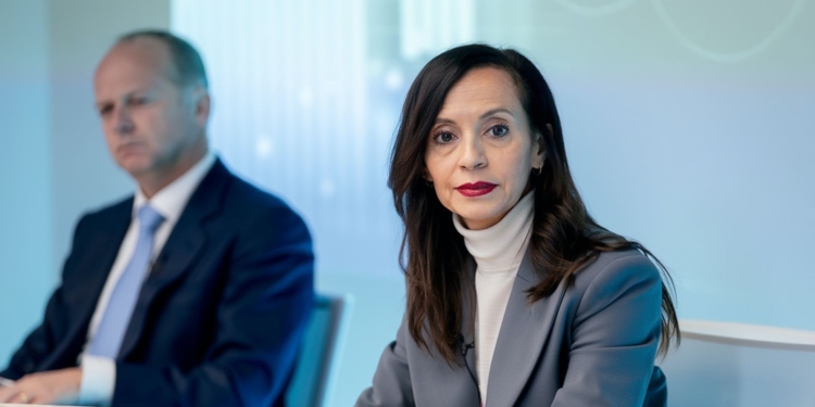 ¿607 millones más? El nuevo sablazo en tu factura de luz. Beatriz Corredor con traje gris en una reunión mientras un hombre aparece desenfocado al fondo.
