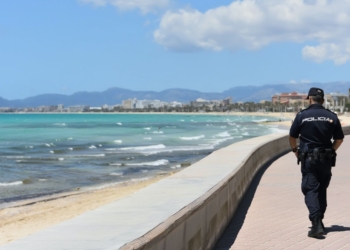 Detenido marroquí en playa de Palma por exhibicionismo ante menores. Un agente de policía camina por un paseo marítimo junto a la playa con el mar y la costa al fondo.