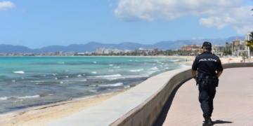 Detenido marroquí en playa de Palma por exhibicionismo ante menores. Un agente de policía camina por un paseo marítimo junto a la playa con el mar y la costa al fondo.