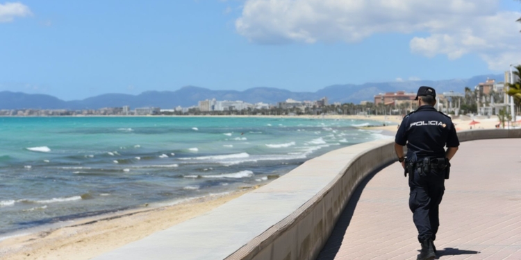 Detenido marroquí en playa de Palma por exhibicionismo ante menores. Un agente de policía camina por un paseo marítimo junto a la playa con el mar y la costa al fondo.