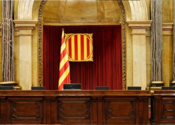 El TSJC impone la bandera española en el Parlament. Sala institucional con una bandera de franjas rojas y amarillas y un escudo sobre un fondo de cortinas rojas.