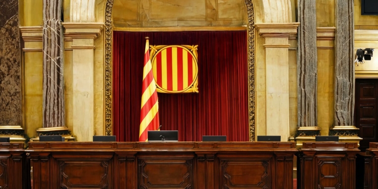 El TSJC impone la bandera española en el Parlament. Sala institucional con una bandera de franjas rojas y amarillas y un escudo sobre un fondo de cortinas rojas.