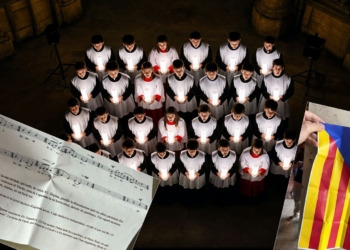 Intento de boicot independentista: expulsados 600 cantantes por querer imponer "Els Segadors" y esteladas en la Sagrada Familia. Coro cantando con velas en un templo, con partitura musical en catalán y una bandera estelada superpuesta.