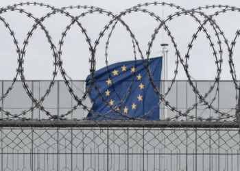 Nueva reglamentación de control de la inmigración en la UE. Alambre de púas en primer plano con la bandera de la Unión Europea ondeando al fondo.