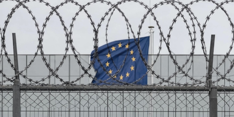 Nueva reglamentación de control de la inmigración en la UE. Alambre de púas en primer plano con la bandera de la Unión Europea ondeando al fondo.