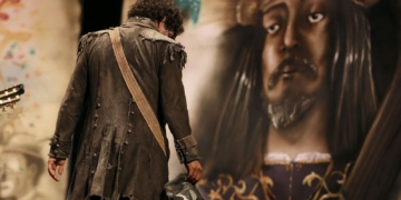 En Cádiz ya se asusta a los niños con "Que viene Juan Carlos". Hombre con traje antiguo y harapiento de espaldas frente a una gran estatua o imagen de un rey coronado.