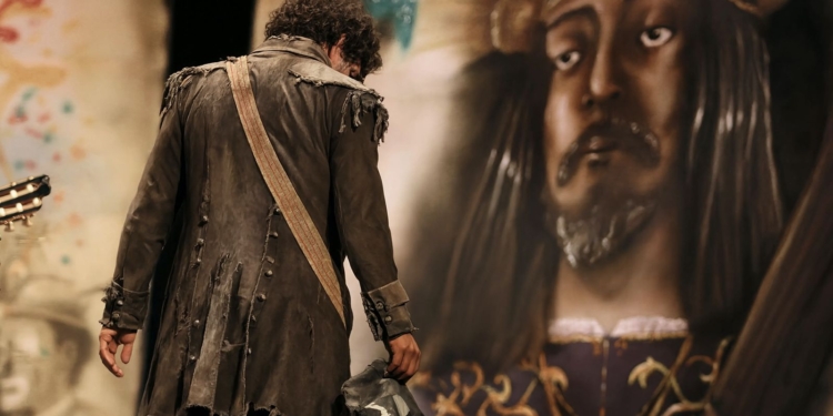 En Cádiz ya se asusta a los niños con "Que viene Juan Carlos". Hombre con traje antiguo y harapiento de espaldas frente a una gran estatua o imagen de un rey coronado.